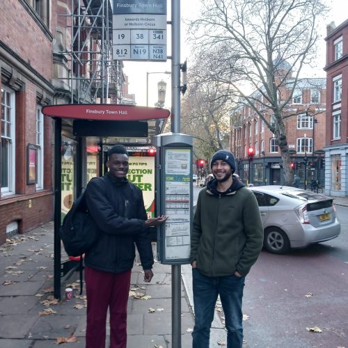 two young men smiling next to a bus stop. One of them is pointing to the timetable.