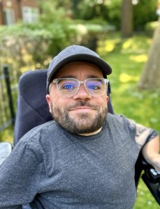 man smiling, sitting in a wheelchair in grassy surroundings wearing a cap and glasses