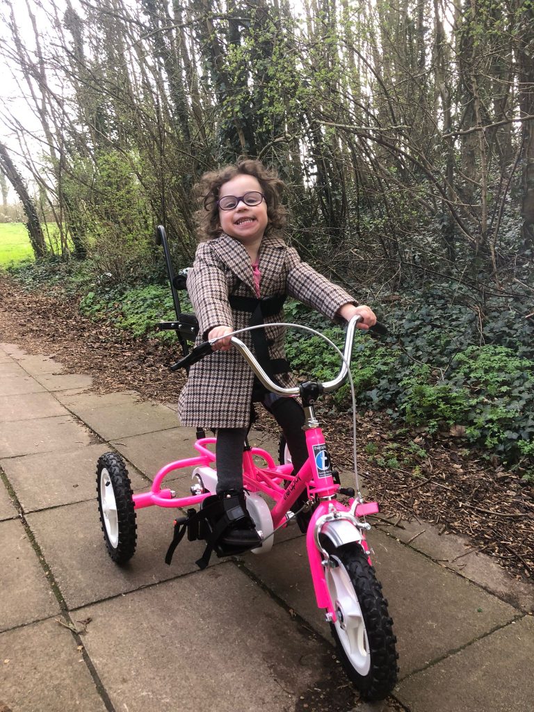 girl in wintery coat on a pink trike smiling in a park